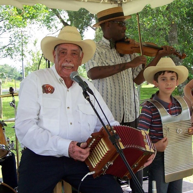 Photo of man on a stage playing music. Young child stands by, also playing music. 