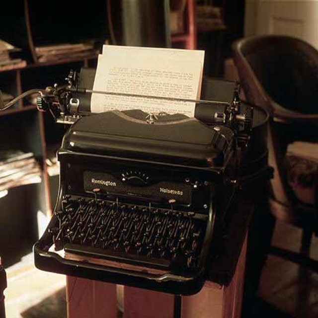 A sepia toned photograph of an old typewriter with paper loaded sits in a office room