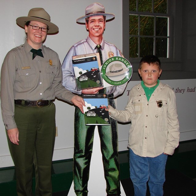 A young boy stands by a real ranger and a cut out ranger as he earns a Jr. Ranger badge