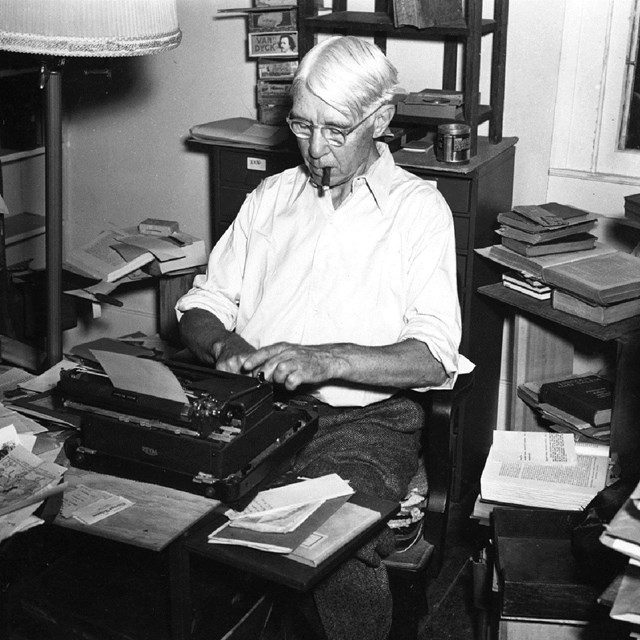 Black and white photo of man  sitting at a typewriter surrounded by books and papers in