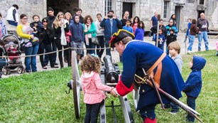 A group of people watch a young visitor being instructed by a ranger dressed as a 1740s soldier.