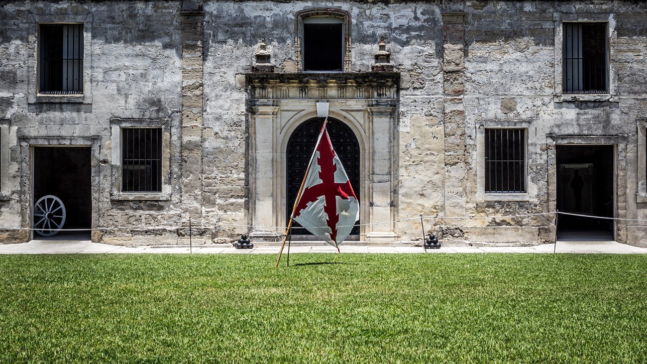 Grass in foreground, splanish flag centered in image, chapel door and walls in background