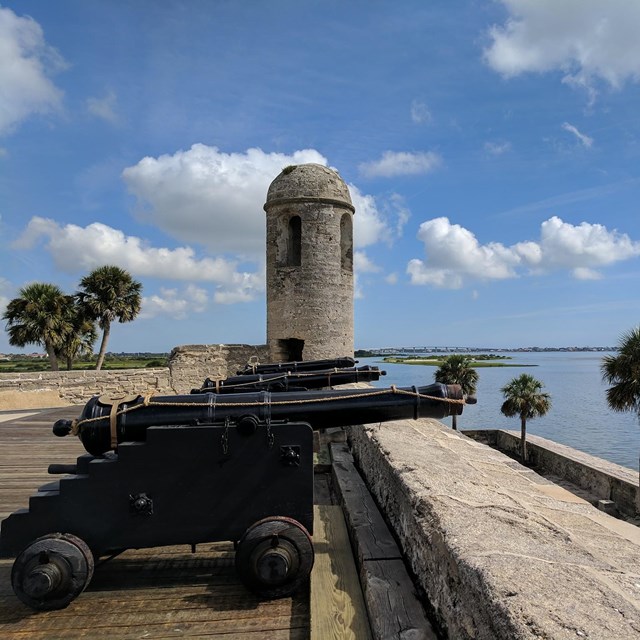 Image of gun deck with cannons, bell tower, water, and palm trees. 