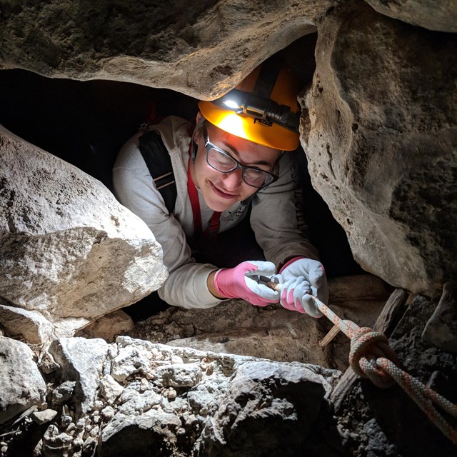 A caver with helmet and headlamp peaks through a small opening in a cave