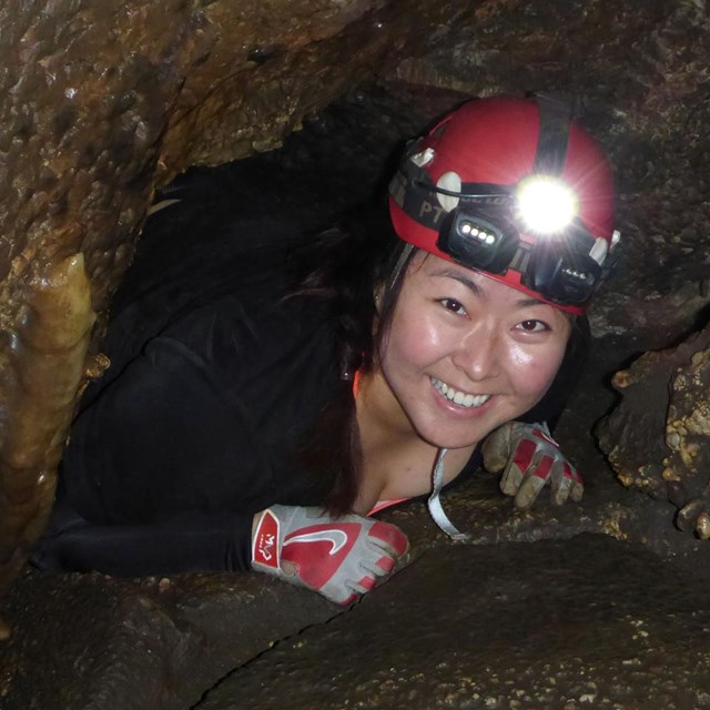 a smiling young woman wearing a helmet, headlamp and gloves climbs through a small opening in a cave