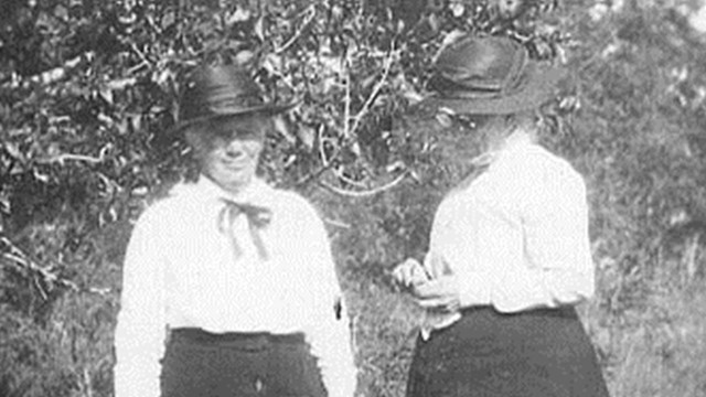 Historic photo of two women wearing hats and long black dresses standing in the grass. 