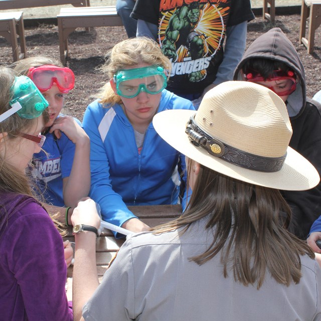 A park ranger has the attention of young students wearing goggles and sitting at a table in a circle