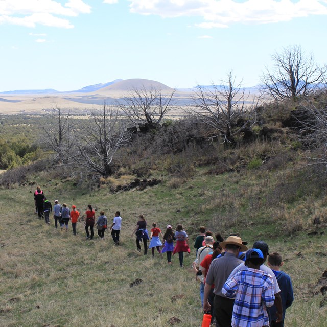 A long line of students walks a trail below a hillside.  Clouds dot a blue sky in the distance.