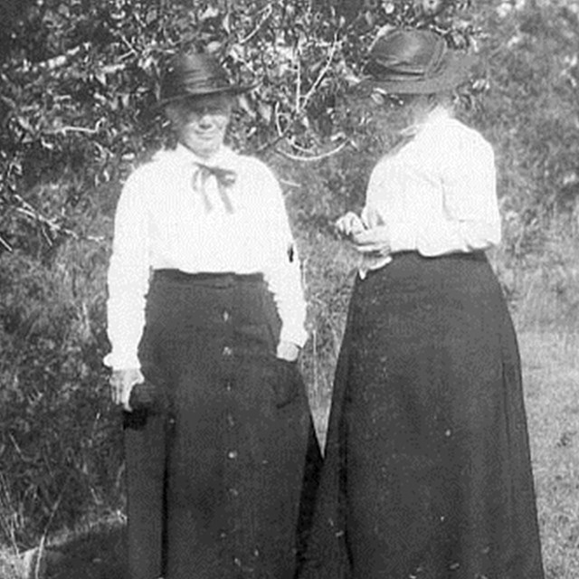 Historic photo of two women wearing hats and long black dresses standing in the grass. 