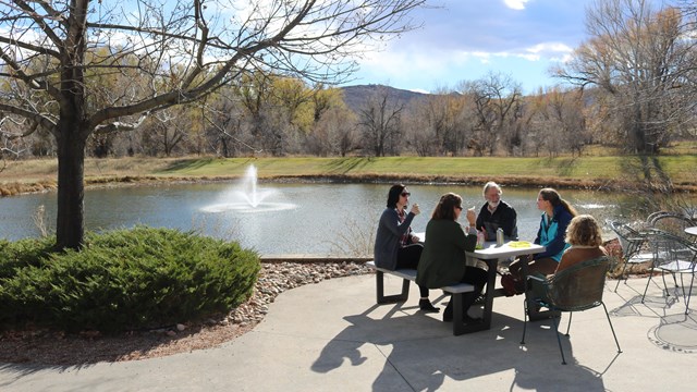 Five people sitting at picnic table with pond and fountain in background on a sunny day