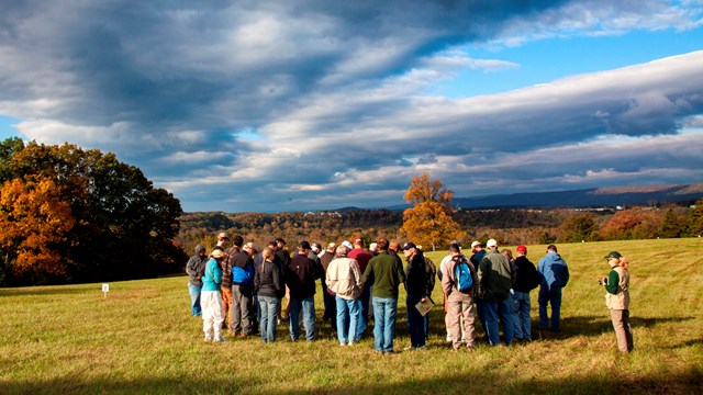 A group of about twenty people stand in a mowed hay field under a blue fall sky.