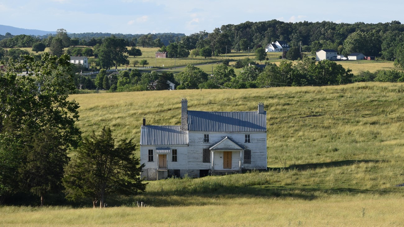 A white two-story farmhouse in a grassy filed with rolling hills in the distance. 