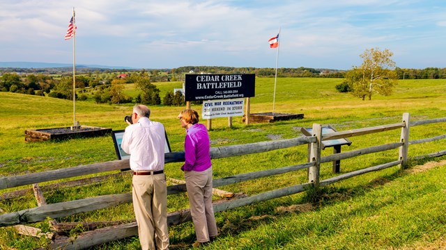 A couple stands at a fence by a historic marker taking pictures of the landscape.