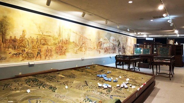 A museum exhibit has a battlefield diorama and a mural painting. 