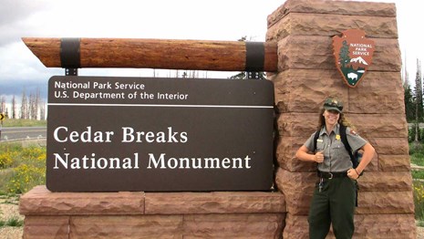 Park ranger standing in front of stone and wood sign for Cedar Breaks National Monument.