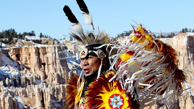 Close up of older native american man with ceremonial dress & orange cliffs in the background. 