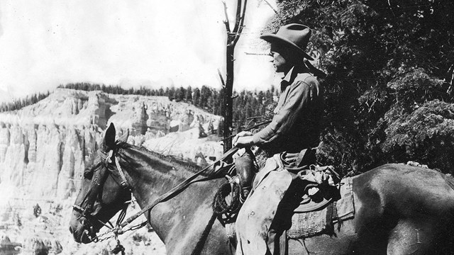 Black and white photo of a man on horseback, looking out at a rocky landscape. 