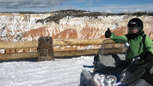 A snowmobiler gives a thumbs up at a snow-covered overlook.
