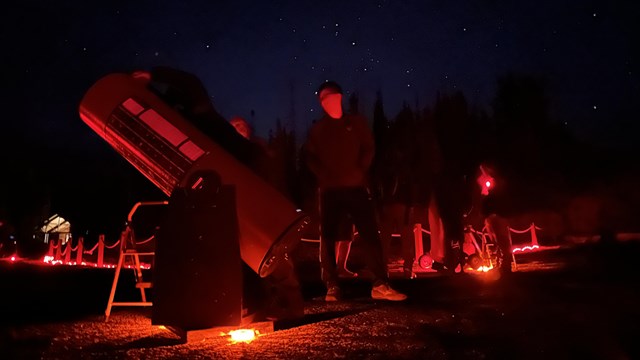 A person stands next to a telescope illuminated by red light before a starry dark sky.