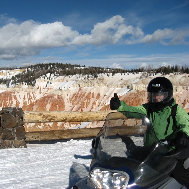 A snowmobiler gives a thumbs up at a snow-covered overlook.