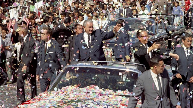 Presidents stand in open car and wave to crowd lining street. Confetti fills the air.