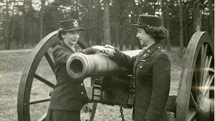 Two female soldiers shake hands over a cannon