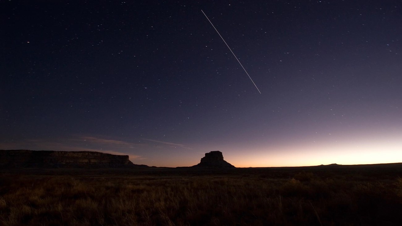 The silhouette of Fajada Butte under a purple and pink night sky with a shooting star above.