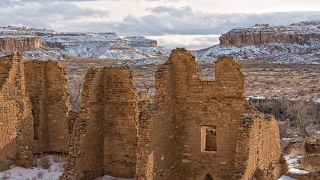 The remnants of a large sandstone structure among an expansive tan canyon covered in snow.