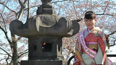 A Japanese woman in cultural attire stands in front of a Japanese stone lantern