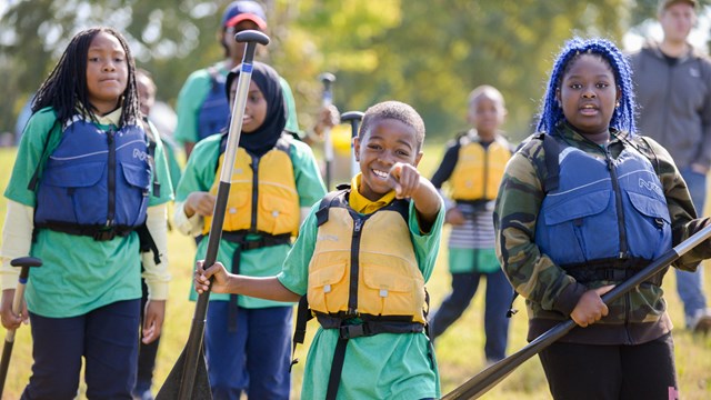 Several young people wearing lifejackets and carrying canoe paddles.