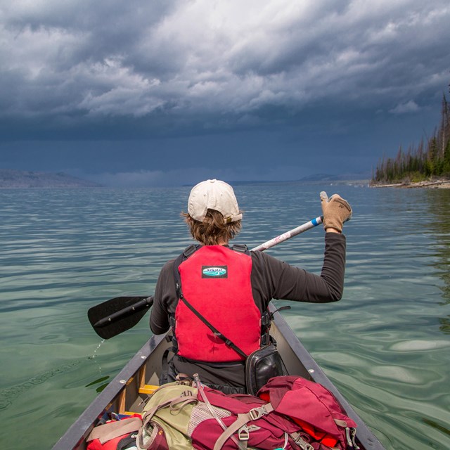 a boy in a red lifejacket paddles a canoe on a lake