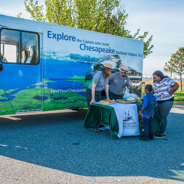 A family and a ranger review documents on a table in front of a bread truck with Chesapeake graphics