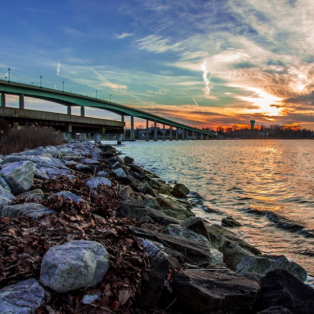 The sun sets behind clouds over a waterway crossed by a concrete bridge.