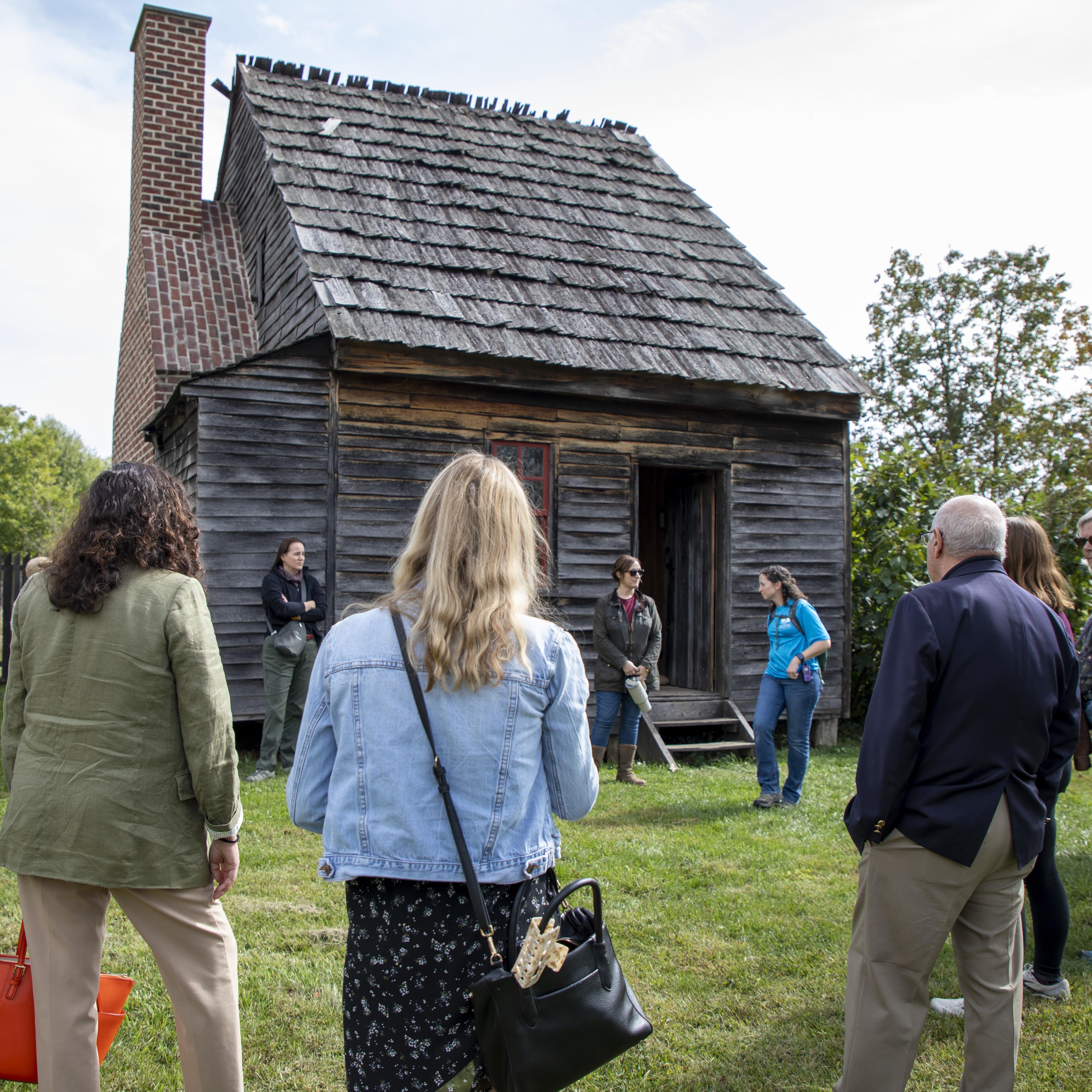 A group of people gathers in front of a small weathered wooden house, listening to a woman speaking.