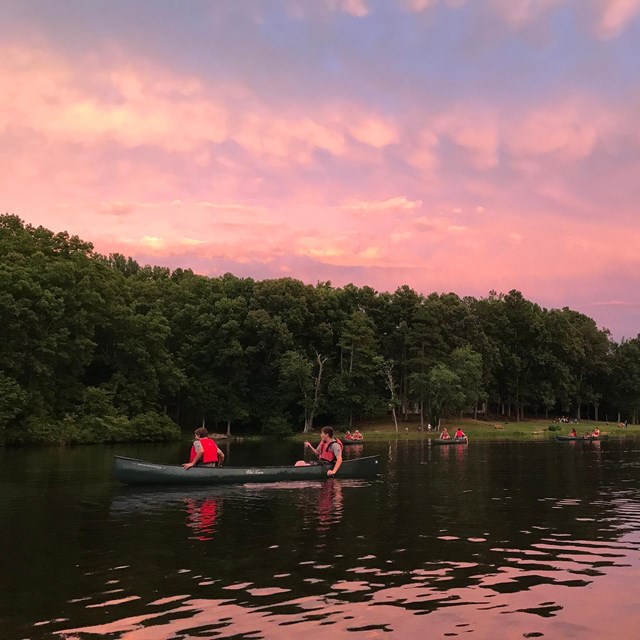 A group paddles canoes under a pink sky at dusk.
