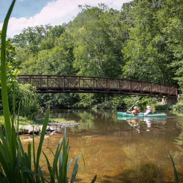 Two people kayak a tandem kayak under a small walking bridge.