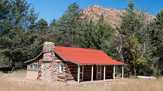 Log cabin in grassy field in front of large rocky hill. 