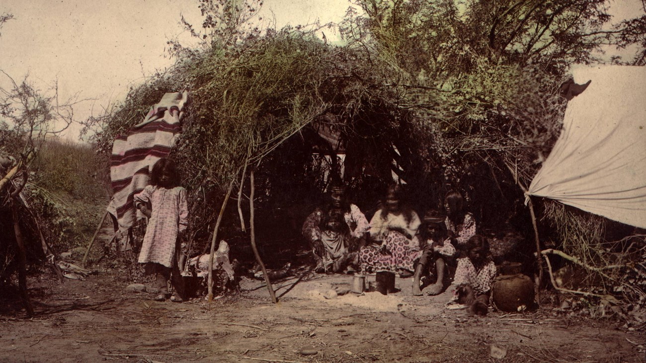 Hand-tinted photograph of wikiup, with adults and children standing outside or sitting inside. 