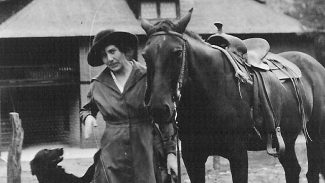 Black and white portrait of woman with horse and dog standing in front of a large house. 