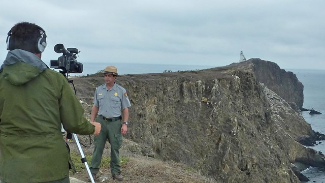 Ranger with video camera filming ranger with lighthouse in background. Credit: Staci Kaye-Carr