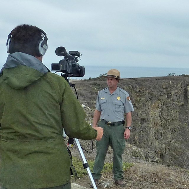Ranger with video camera filming ranger with lighthouse in background. Credit: Staci Kaye-Carr