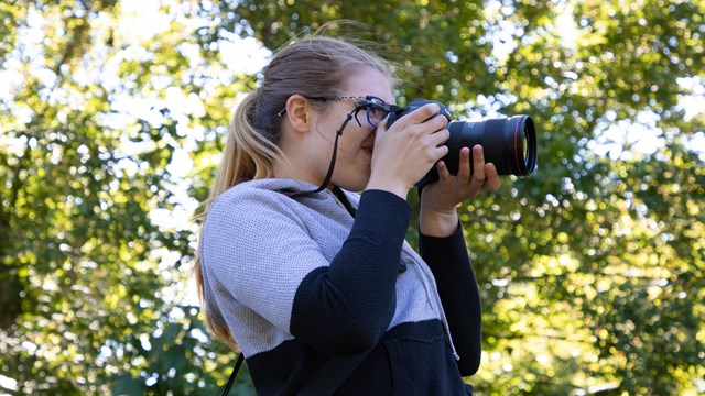 Young woman with blonde hair in a ponytail using a camera to take a photograph.