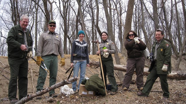 Six people standing in a row in the woods with tools.