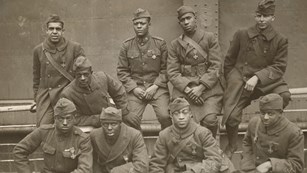 Black and white photo of nine men in two rows shoulder to shoulder wearing WW1 uniforms
