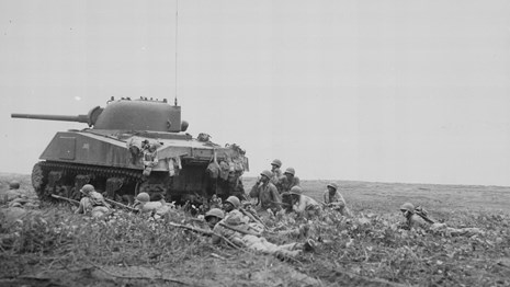 Black and white photo of a army tank on the left side of photo protecting soldiers laying prone.