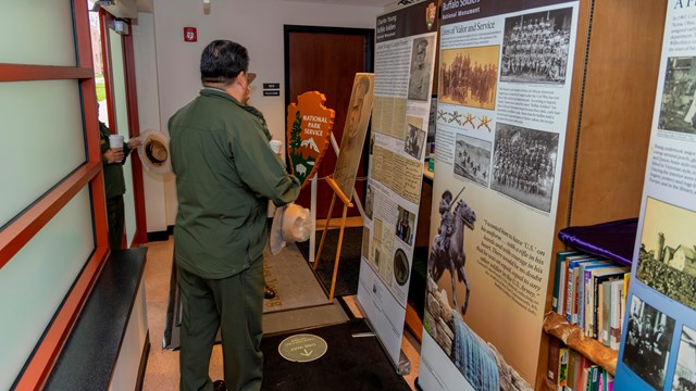 A man in a green jacket stands in front of several banners with information and photos on them.