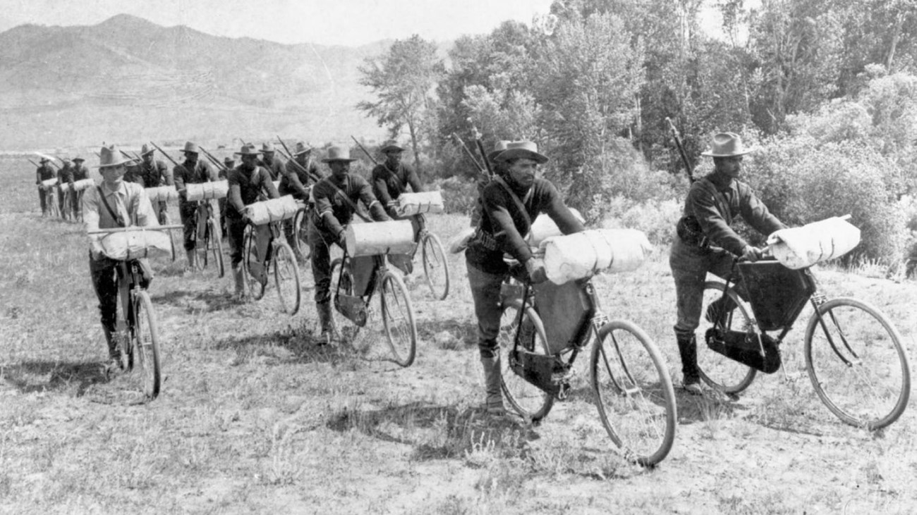 Black and white photo of two columns of African American army soldiers riding bikes towards the view