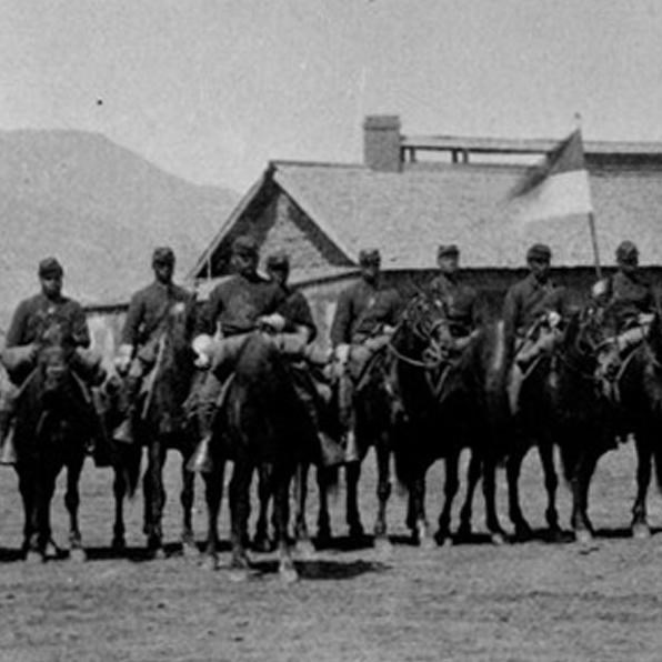 Several Black soldiers mounted on horses aligned in a row in front of a 1-story building