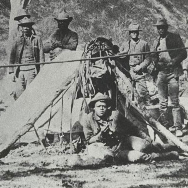 Black soldiers laying down in front of a tent with several more standing behind the tent
