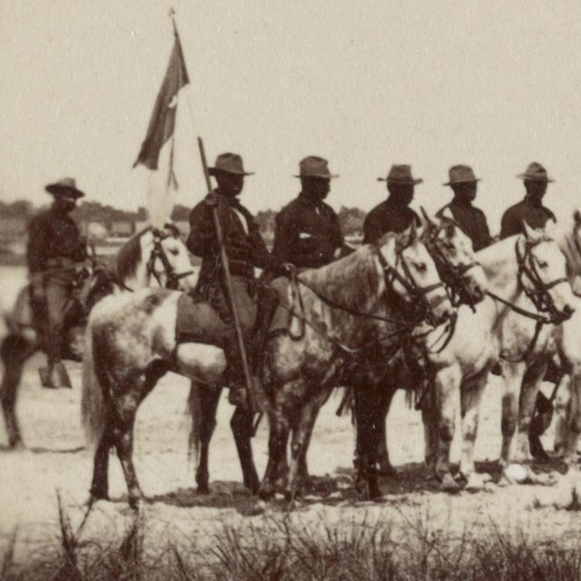 Several Black soldiers sitting atop horses and a couple holding flags on poles, in formation